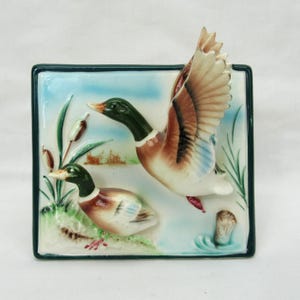 Könnte beinhalten: Keramik-Wandbild mit zwei Stockenten in einer Feuchtlandschaft. Eine Ente ist im Flug mit ausgebreiteten Flügeln, die andere ruht am Ufer. Die Platte hat einen grünen Rand und einen hellblauen Hintergrund.