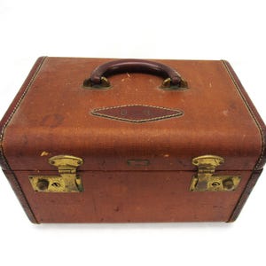 May include: A rectangular brown travel case with a dark brown leather handle and brass hardware. The top features a diamond-shaped leather patch with the embossed letters D.S.H. The case has two brass latches on the front and visible wear.
