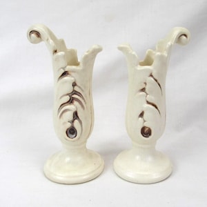 May include: A pair of off-white ceramic vases featuring a tall, slender body with a flared, scrolled rim. Each vase displays a raised, leaf-like relief pattern along the front, accented with brown detailing that highlights the textured design.