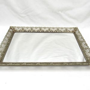 May include: A rectangular silver-toned vanity tray with a mirrored surface. The tray has an ornate, openwork filigree border. The design is symmetrical, with a repeating pattern along the edges. The tray is likely made of metal and is suitable for displaying jewelry or perfumes.