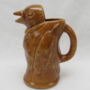 May include: A brown ceramic pitcher in the shape of a bird. The pitcher has a handle and a spout that resembles a bird's beak. The bird is wearing a suit. The pitcher is likely vintage.