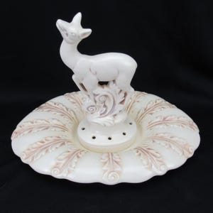 Puede incluir: Una figura de ciervo de cerámica blanca sobre un plato decorativo con volantes. El plato presenta un patrón en relieve en forma de hoja en un tono rosa apagado. El ciervo está sobre una base decorativa con varios agujeros pequeños.