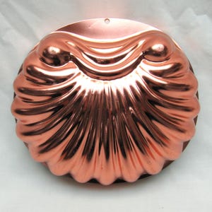 May include: A copper-colored, shell-shaped mold with a scalloped edge. The mold has a detailed, textured surface and a small hole at the top for hanging. The design features a series of curved ridges radiating from the center.
