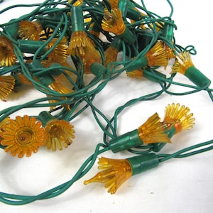 May include: A string of decorative lights featuring translucent amber-colored plastic covers shaped like small, multi-petaled flowers. The flower-shaped covers are attached to a dark green electrical cord, creating a tangled cluster of lights.