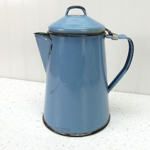 May include: A vintage blue enamelware coffee pot with a lid and handle. The pot has a spout for pouring and a dark rim. The enamel is chipped in places, giving it a rustic appearance. The pot is a light blue color.