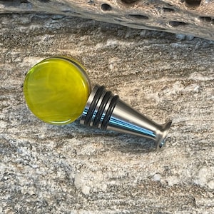 May include: A decorative bottle stopper with a stainless steel base and a round, yellow-green glass top. The glass has a swirled pattern. The stopper is designed to fit into a bottle.