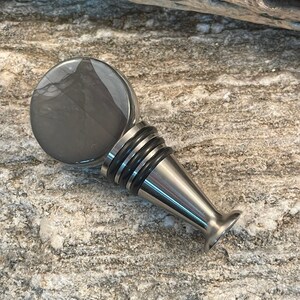 May include: A silver bottle stopper with a round, gray, marbled top. The stopper has a black rubber ring around the top and a silver base.