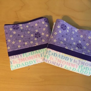 May include: Two purple fabric pouches with a white floral pattern. The pouches have a purple ribbon and a white background with the text "I love Mommy I love Daddy" repeated.