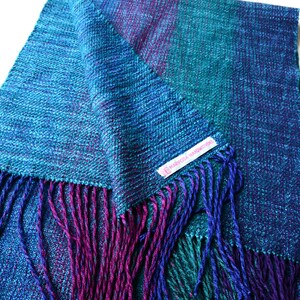 Op de afbeelding: Een handgeweven sjaal in tinten blauw, teal en paars. De sjaal heeft een getextureerde weefsel en gefranjerde randen in complementaire kleuren. Een klein label met tekst is zichtbaar.