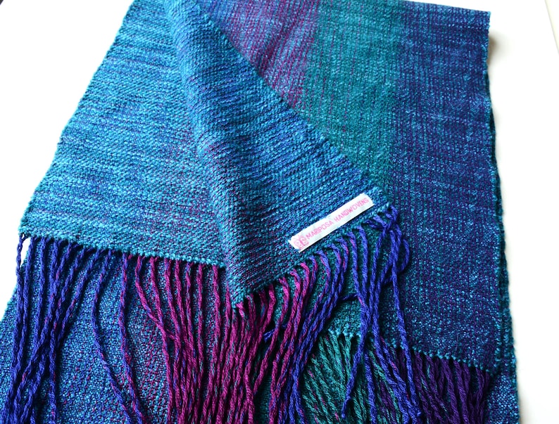 Op de afbeelding: Een handgeweven sjaal in tinten blauw, teal en paars. De sjaal heeft een getextureerde weefsel en gefranjerde randen in complementaire kleuren. Een klein label met tekst is zichtbaar.