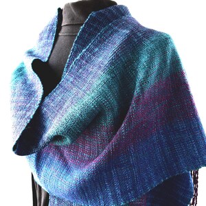 Op de afbeelding: Een handgeweven sjaal in tinten blauw, blauwgroen en paars. Het textiel is gedrapeerd over een zwarte vorm, waarbij de ingewikkelde weefsel en kleurovergangen worden getoond. De sjaal heeft een gefranjerde rand en een getextureerde uitstraling, geschikt voor koel weer.
