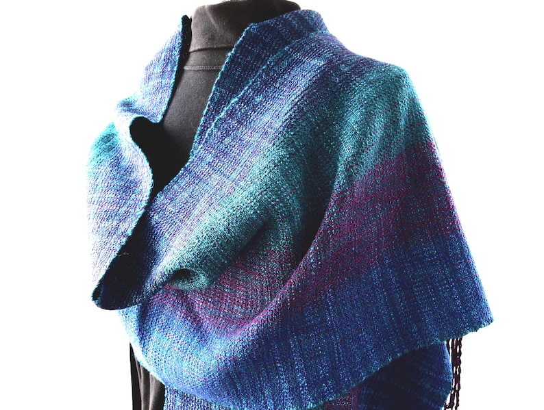 Op de afbeelding: Een handgeweven sjaal in tinten blauw, blauwgroen en paars. Het textiel is gedrapeerd over een zwarte vorm, waarbij de ingewikkelde weefsel en kleurovergangen worden getoond. De sjaal heeft een gefranjerde rand en een getextureerde uitstraling, geschikt voor koel weer.