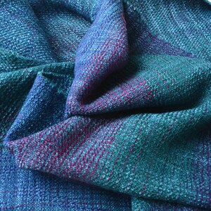 Op de afbeelding: Een handgeweven sjaal in tinten van blauwgroen, blauw en paars. De stof heeft een getextureerde uitstraling met zichtbare weefdetails. De sjaal is gedrapeerd, wat het samenspel van kleuren en de drapering van de stof laat zien.