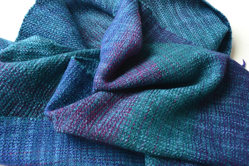 Op de afbeelding: Een handgeweven sjaal in tinten van blauwgroen, blauw en paars. De stof heeft een getextureerde uitstraling met zichtbare weefdetails. De sjaal is gedrapeerd, wat het samenspel van kleuren en de drapering van de stof laat zien.