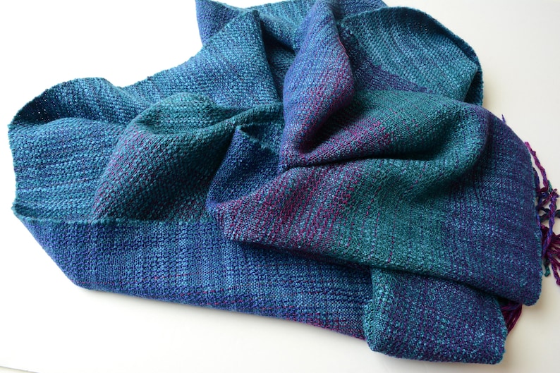 Op de afbeelding: Een handgeweven sjaal in tinten blauw, blauwgroen en paars. De textuurstof is gedrapeerd en toont de kleurvariaties en het geweven patroon. De sjaal heeft een gefranjerde rand.