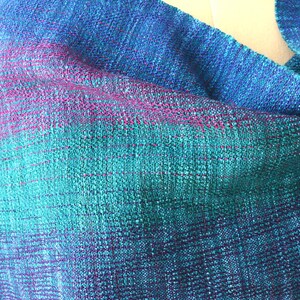 Op de afbeelding: Een handgeweven sjaal in tinten blauw, teal en paars. De stof heeft een textuur, met een gradi&euml;nteffect dat overgaat van diepblauw naar teal en vervolgens naar paars. De sjaal is gedrapeerd en toont het geweven patroon.