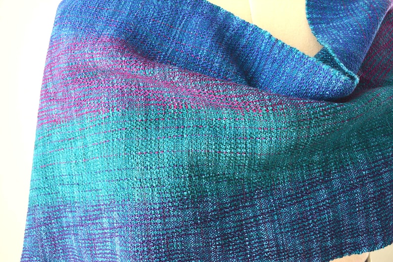 Op de afbeelding: Een handgeweven sjaal in tinten blauw, teal en paars. De stof heeft een textuur, met een gradi&euml;nteffect dat overgaat van diepblauw naar teal en vervolgens naar paars. De sjaal is gedrapeerd en toont het geweven patroon.
