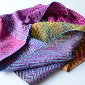 May include: A colorful, woven scarf with a chevron pattern. The scarf features a gradient of colors, including pink, purple, yellow, and teal. The edges are finished with a contrasting teal border. This accessory is suitable for various occasions.