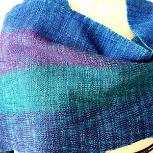 Op de afbeelding: Een handgeweven sjaal in tinten blauw, teal en paars. De stof heeft een textuur, met een subtiel gradi&euml;nteffect. De sjaal is over een mannequin gedrapeerd, wat de vorm en de drape laat zien.