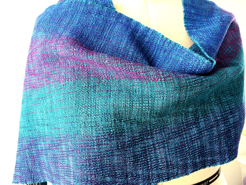 Op de afbeelding: Een handgeweven sjaal in tinten blauw, teal en paars. De stof heeft een textuur, met een subtiel gradi&euml;nteffect. De sjaal is over een mannequin gedrapeerd, wat de vorm en de drape laat zien.