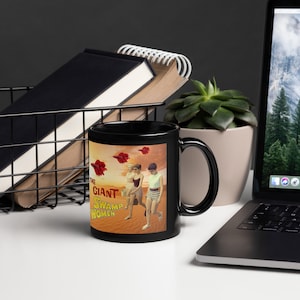 Könnte beinhalten: Schwarze Kaffeetasse mit einem Grafikdruck von drei Frauen, die an einem Strand mit roten fliegenden Fischen am Himmel entlanglaufen. Der Text "The Giant Swamp Women" ist auf die Tasse gedruckt.