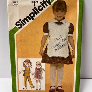 May include: Vintage Simplicity sewing pattern for a child's dress and apron, size 3. The pattern features a drawing of a young girl wearing the dress and apron. The pattern includes the text "PAID Hancock Fab Paducah, K".