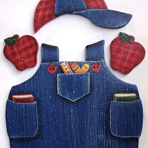 May include: A blue denim overall with two pockets, one with a red book labeled "MATH" and the other with a green book labeled "A B C's". The overall has a red and blue plaid baseball cap, two red apples, and a ruler and pencil in the front pocket.