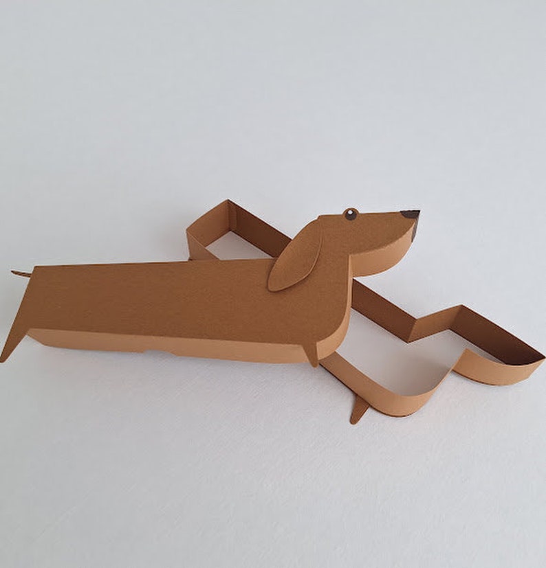 May include: A brown paper dachshund shaped box with a cut-out design. The box is open and the inside is white.
