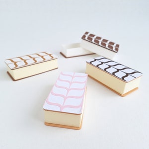 May include: Four decorative boxes with different patterns. The boxes are white with a brown, black, or pink design. The boxes are all closed and are sitting on a white surface.