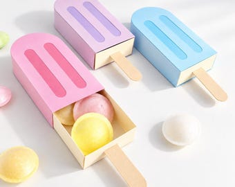 May include: Three popsicle-shaped cardboard boxes in pink, purple, and blue, each featuring a wooden stick handle. The pink box is open, revealing round, pastel-colored candies inside, while additional loose candies are scattered nearby.