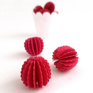 Puede incluir: Esferas decorativas de papel rojo con un diseño de borde festoneado. Varias esferas están dispersas sobre una superficie blanca, con más esferas en un recipiente blanco festoneado en el fondo. Las esferas son de color rojo vibrante.