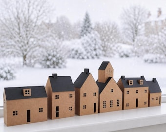 Op de afbeelding: Een rij van zes miniatuurhuisjes gemaakt van bruin karton met zwarte daken en uitgesneden ramen en deuren. De huisjes variëren in hoogte en architecturale vorm, waarbij er één een klok op de voorgevel heeft, opgesteld op een witte vensterbank.