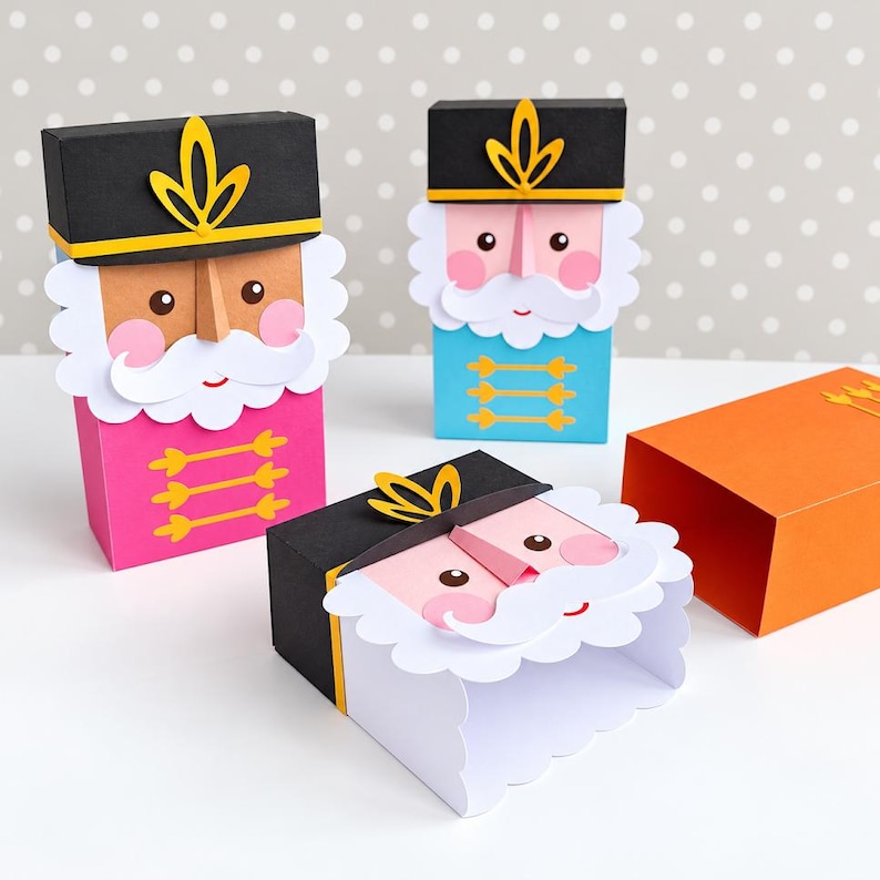 Op de afbeelding: Drie papieren notenkraker-cadeaudoosjes met zwarte hoeden met gele accenten, witte baarden en roze wangen. E&eacute;n doosje is roze, &eacute;&eacute;n is blauw en &eacute;&eacute;n is oranje. De doosjes hebben rechthoekige lichamen en decoratieve gele versieringen aan de voorzijde.