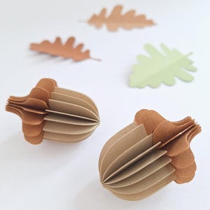 Op de afbeelding: Twee papieren eikels met bruine doppen en bruine lichamen worden getoond. Drie papieren bladeren in bruin- en groentinten zijn op de achtergrond verspreid. De eikels hebben een gelaagd, driedimensionaal ontwerp.