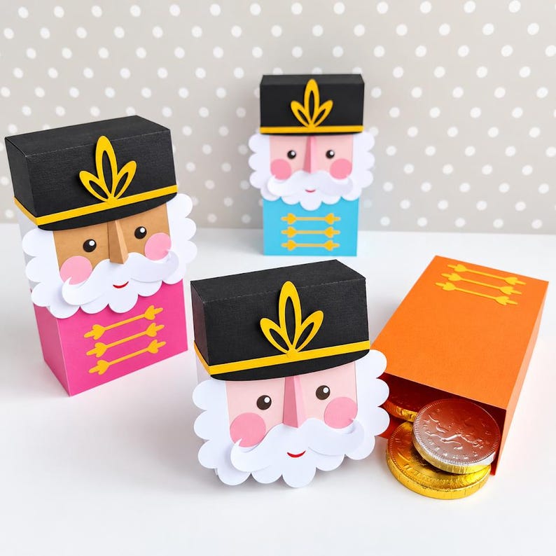 Op de afbeelding: Drie papieren notenkrakerdoosjes met zwarte hoeden, witte baarden en roze wangen. E&eacute;n doosje is roze, &eacute;&eacute;n blauw en &eacute;&eacute;n oranje. Het oranje doosje staat open en toont twee ronde gouden en zilveren chocolademunten in de rechthoekige verpakking.