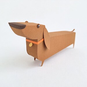 May include: A brown paper dachshund with a orange collar and a gold tag. The dachshund is standing on a white surface.