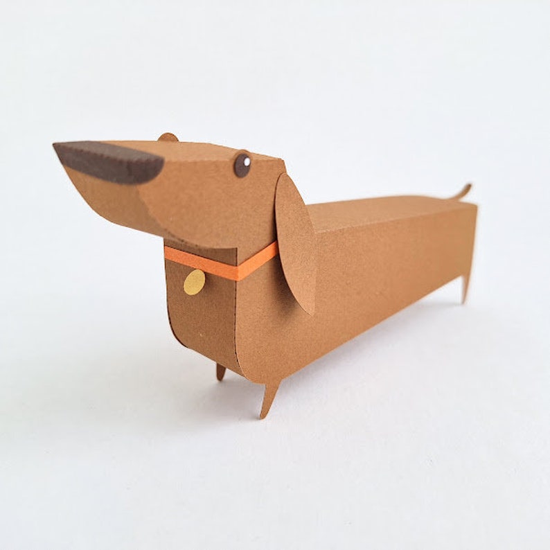 May include: A brown paper dachshund with a orange collar and a gold tag. The dachshund is standing on a white surface.