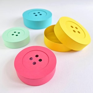 May include: A collection of colorful, button-shaped boxes in shades of pink, yellow, blue, and green. Each box has a lid with four small holes, resembling a button. One yellow box is open, revealing its interior.