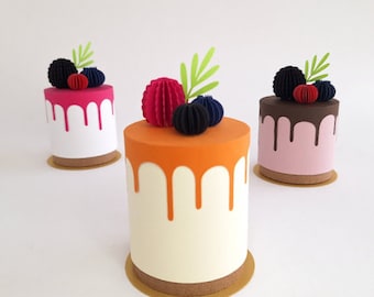 May include: Three miniature paper cakes decorated with colorful paper flowers and berries. The cakes are topped with different colored drizzles: pink, orange, and brown.