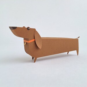May include: A brown paper dachshund with an orange collar. The dachshund is made of paper and has a 3D effect.