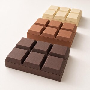 May include: Three rectangular boxes arranged in a row, each featuring a raised, grid-like surface pattern that resembles a chocolate bar. The boxes are colored in shades of dark brown, medium brown, and light beige, with a matte finish.
