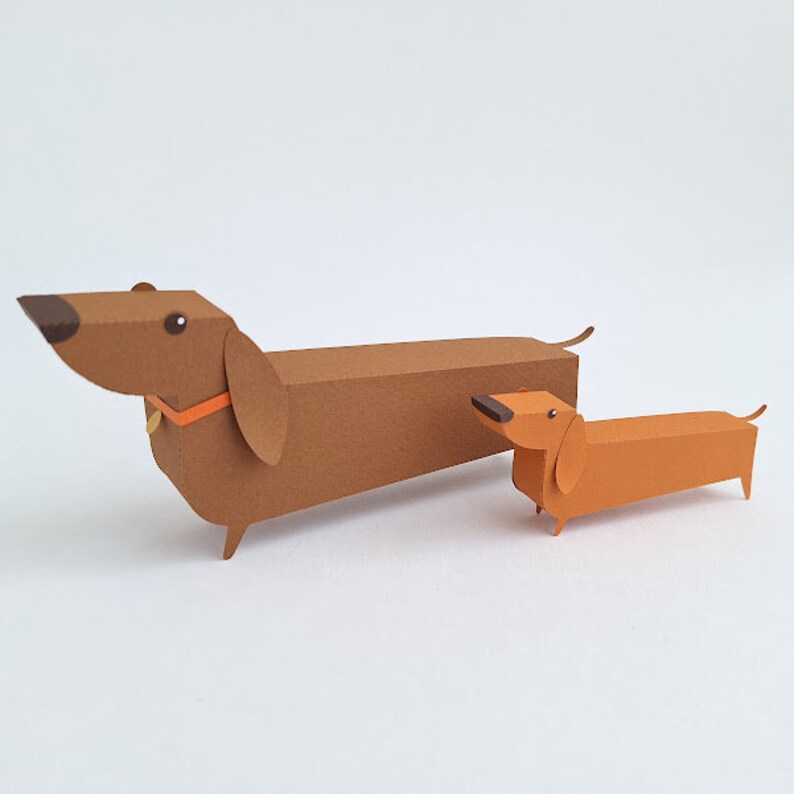 May include: Two paper dachshund dogs, one brown and one orange, are standing side by side on a white surface. The larger dachshund has a brown collar with an orange accent.