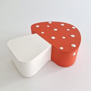 Pode incluir: Uma caixa de papel em forma de cogumelo vermelha e branca com bolinhas brancas. O cogumelo &eacute; feito de papel e tem um caule branco e um chap&eacute;u vermelho.