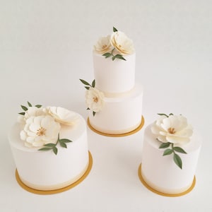 Puede incluir: Tres pasteles blancos de varios niveles decorados con flores de papel blanco y hojas verdes. Los pasteles están sobre bases de pastel doradas.