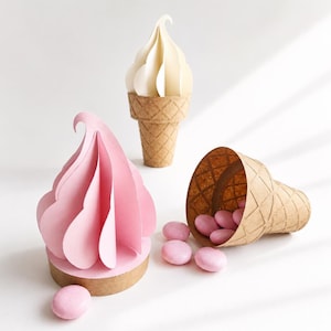 May include: Three-dimensional paper ice cream cones featuring a tall cream-colored cone, a pink soft-serve style paper sculpture on a brown base, and a brown waffle-textured cone tipped over with several small, round, light pink candies.