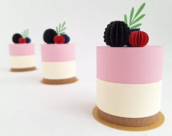May include: Three miniature paper cakes with pink and white frosting, decorated with black and red paper berries and green leaves.