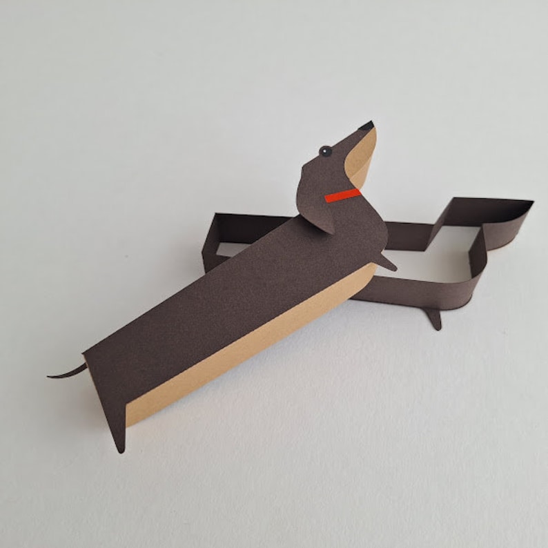 May include: A brown and tan paper dachshund with a red collar, standing on a white surface.
