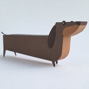 May include: A brown and tan paper dachshund figurine. The dachshund is standing on a white surface.