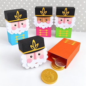 Op de afbeelding: Vier papieren notenkraker-cadeaudoosjes in blauw, roze, groen en oranje, elk met een zwarte hoed met geel embleem, witte baard en roze wangen. Een oranje doosje ligt open en toont gouden en zilveren chocolademunten aan de binnenkant.