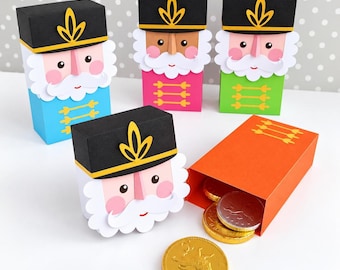 May include: Four paper nutcracker gift boxes in blue, pink, green, and orange, each featuring a black hat with a yellow emblem, white beard, and pink cheeks. An orange box lies open, revealing gold and silver chocolate coins inside.